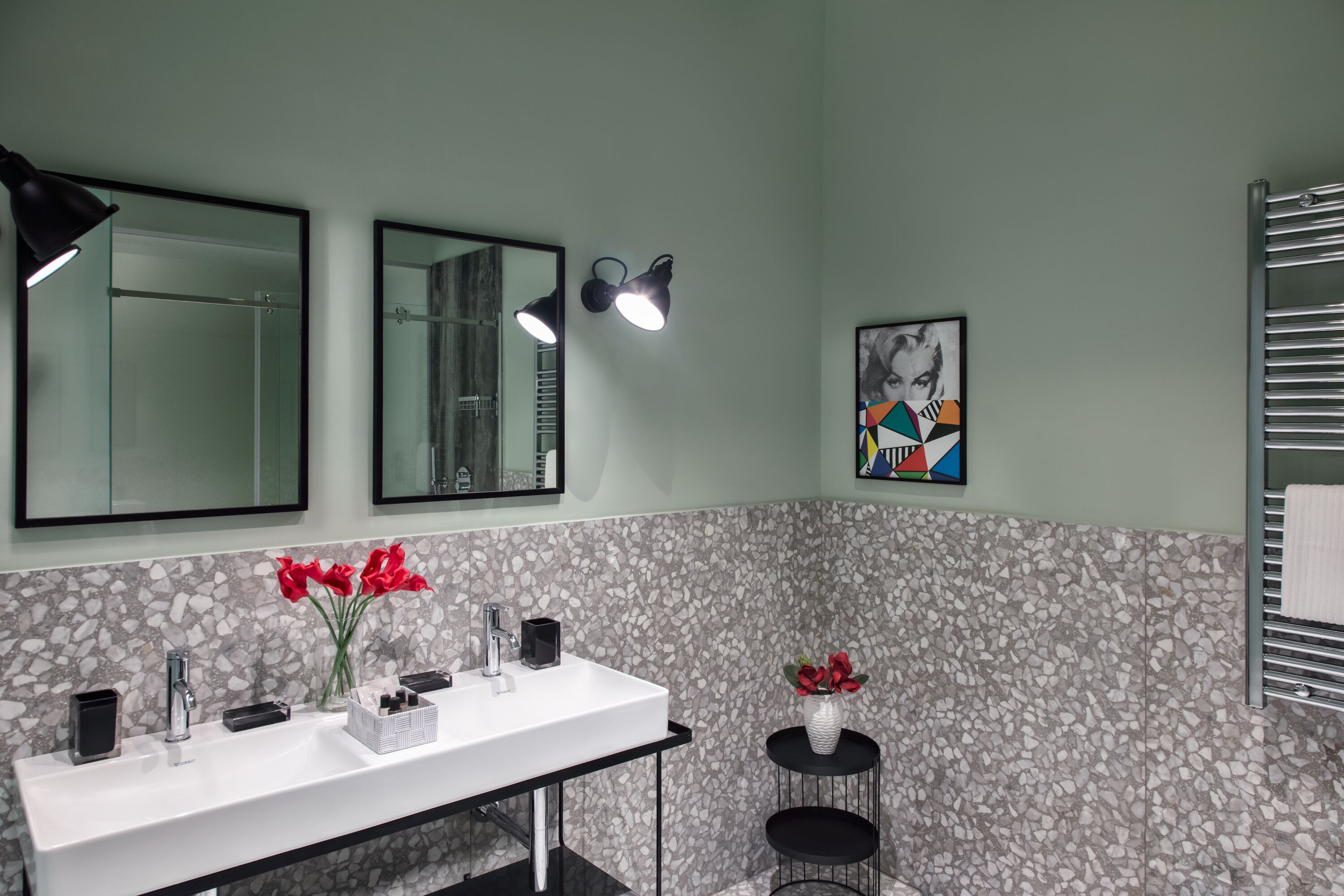 A bathroom with a white sink, two mirrors, a towel rack, and a small round stool with a vase of red flowers.