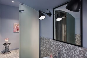Une salle de bain moderne avec une douche à l’italienne en verre, un grand miroir et un vase de fleurs posé sur une petite table.