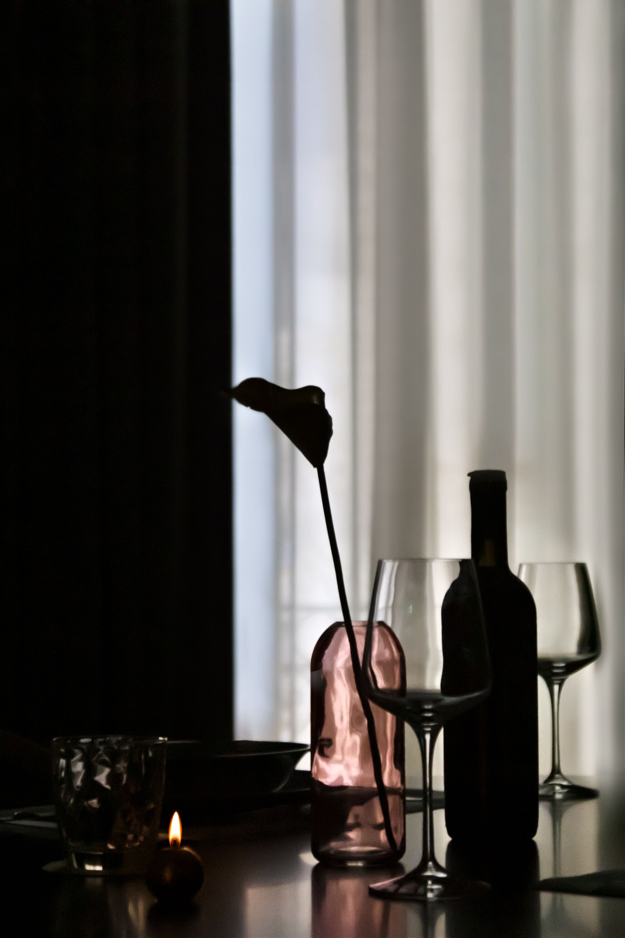 Een kamer in gedimd licht met een kaars, een glazen vaas met een bloem en wijnglazen op een tafel.