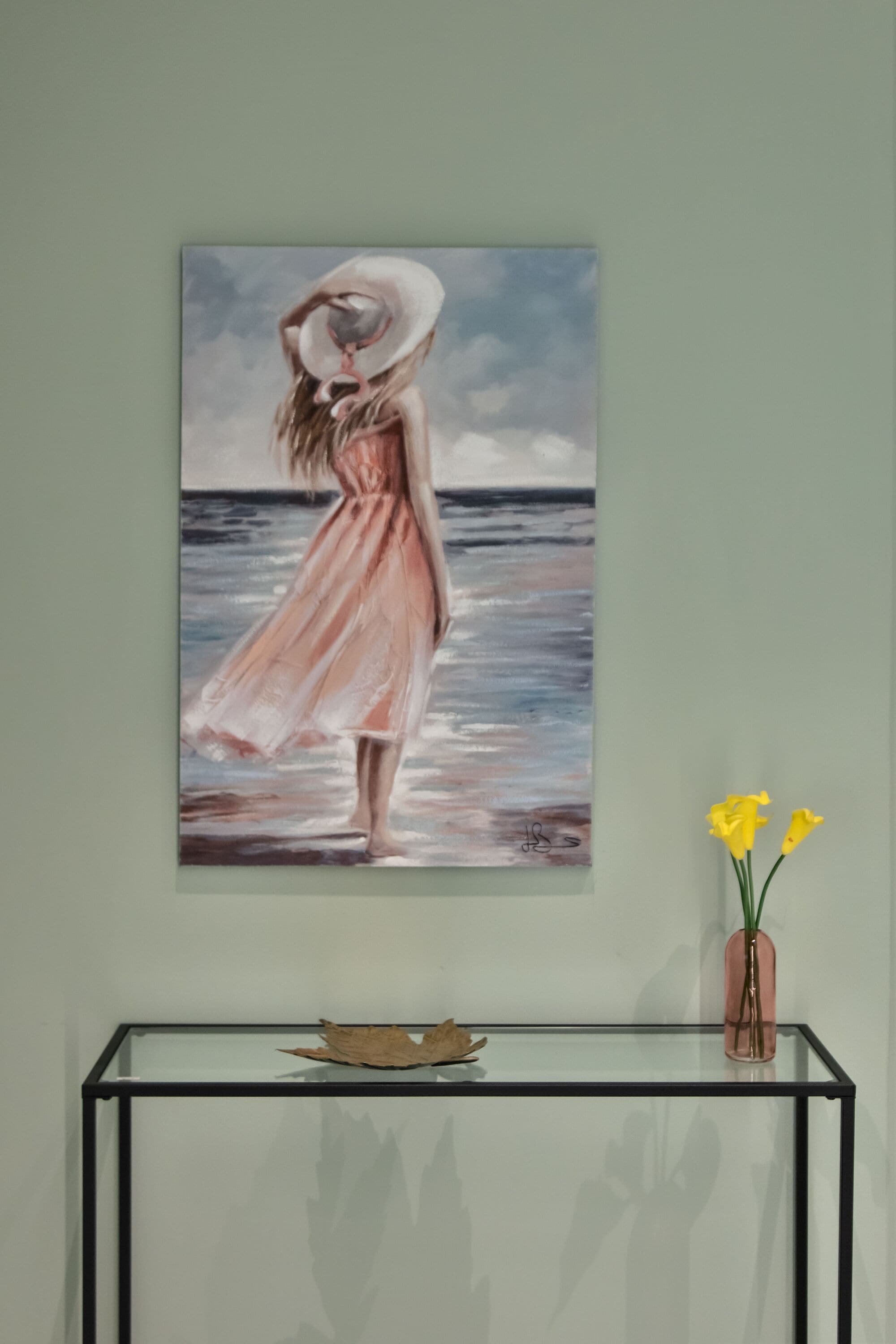 A painting of a woman in a pink dress and hat by the sea, displayed on a wall above a glass table with a vase of yellow flowers.