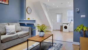 Een moderne woonkamer met een bank, salontafel en een potplant. Een trap leidt naar een keukenruimte met een wasmachine.