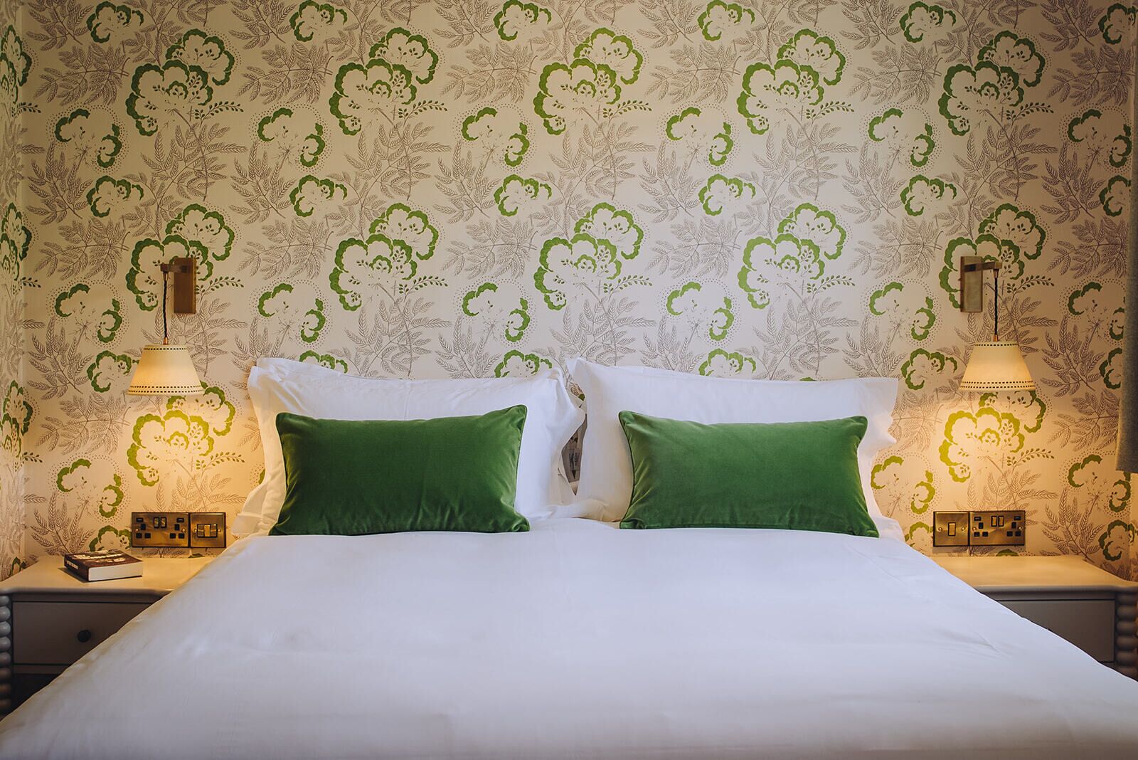 Een bed met witte lakens en twee groene kussens, met aan weerszijden nachtkastjes met lampen en een stekkerdoos.
