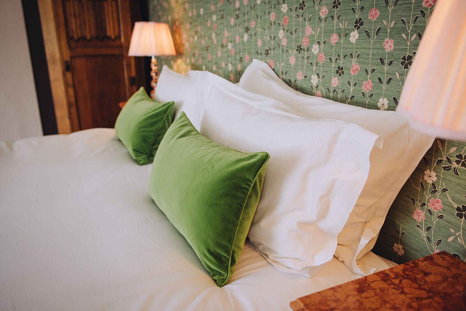 A bed with white linens and green pillows, a wooden headboard with floral wallpaper, and a nightstand with a lamp.