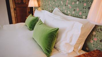 A bed with white linens and green pillows, a wooden headboard with floral wallpaper, and a nightstand with a lamp.