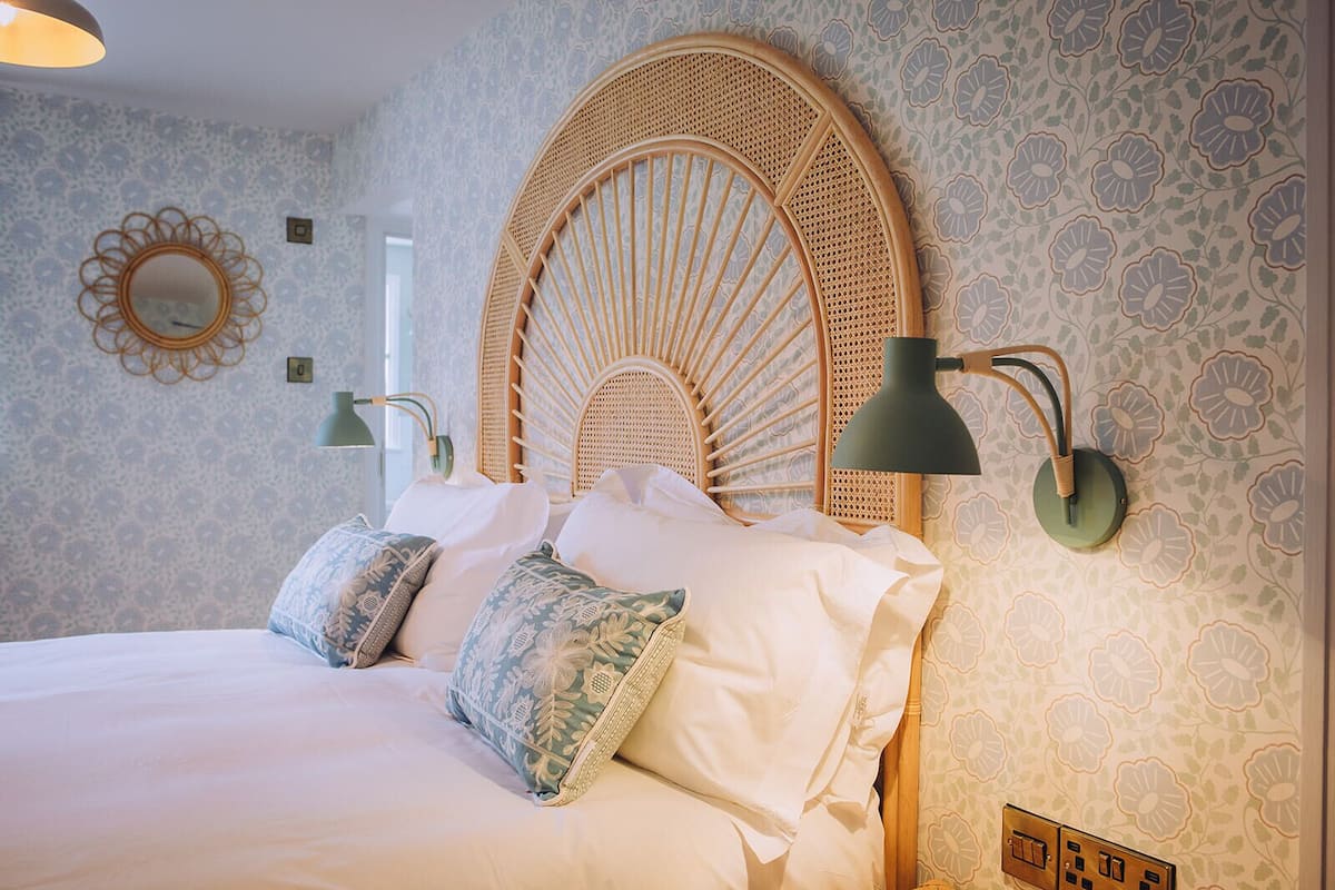 A bed with a rattan headboard, two wall-mounted lamps, and patterned wallpaper.