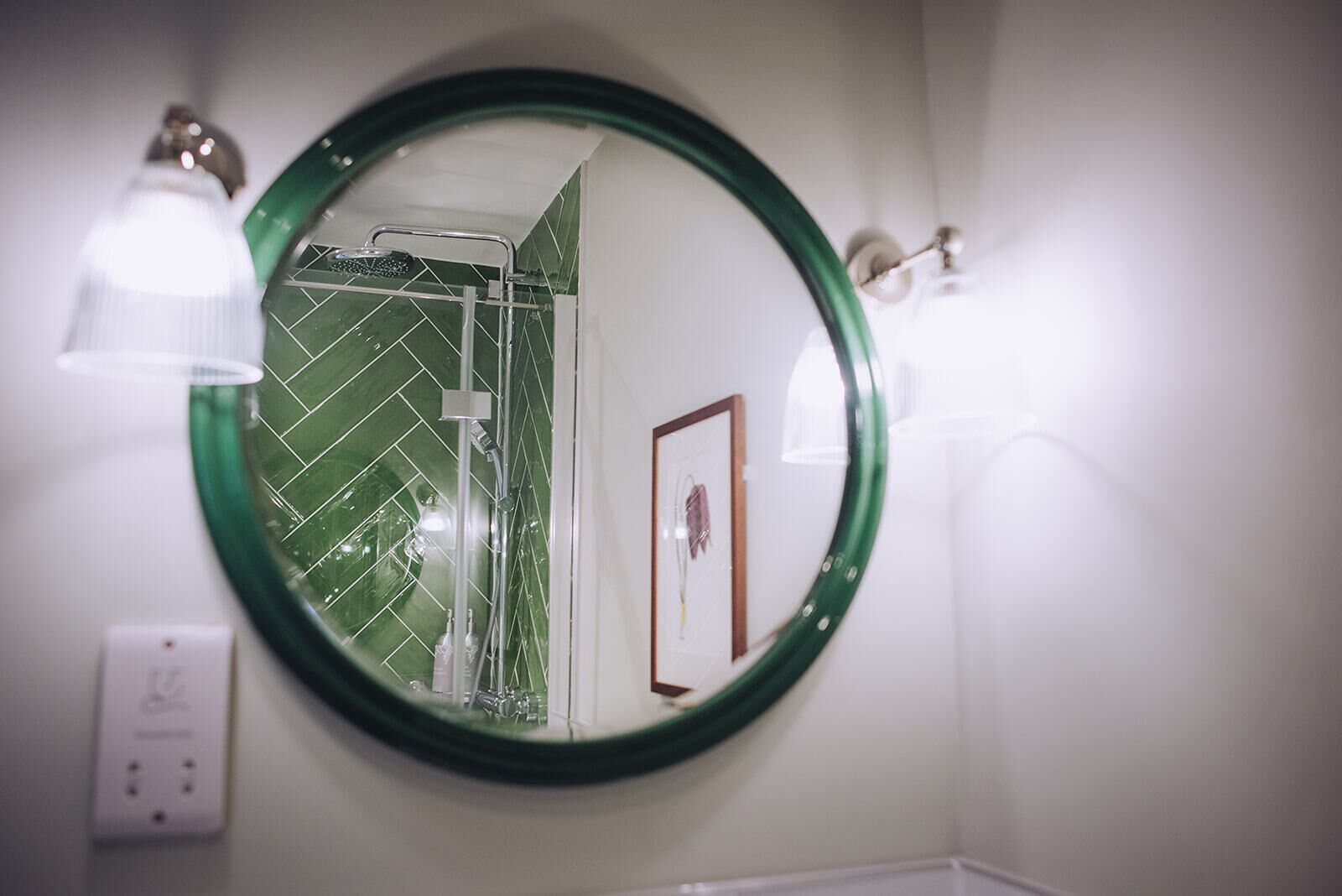 A bathroom with a round mirror, green-tiled walls, a framed picture, and a light fixture.