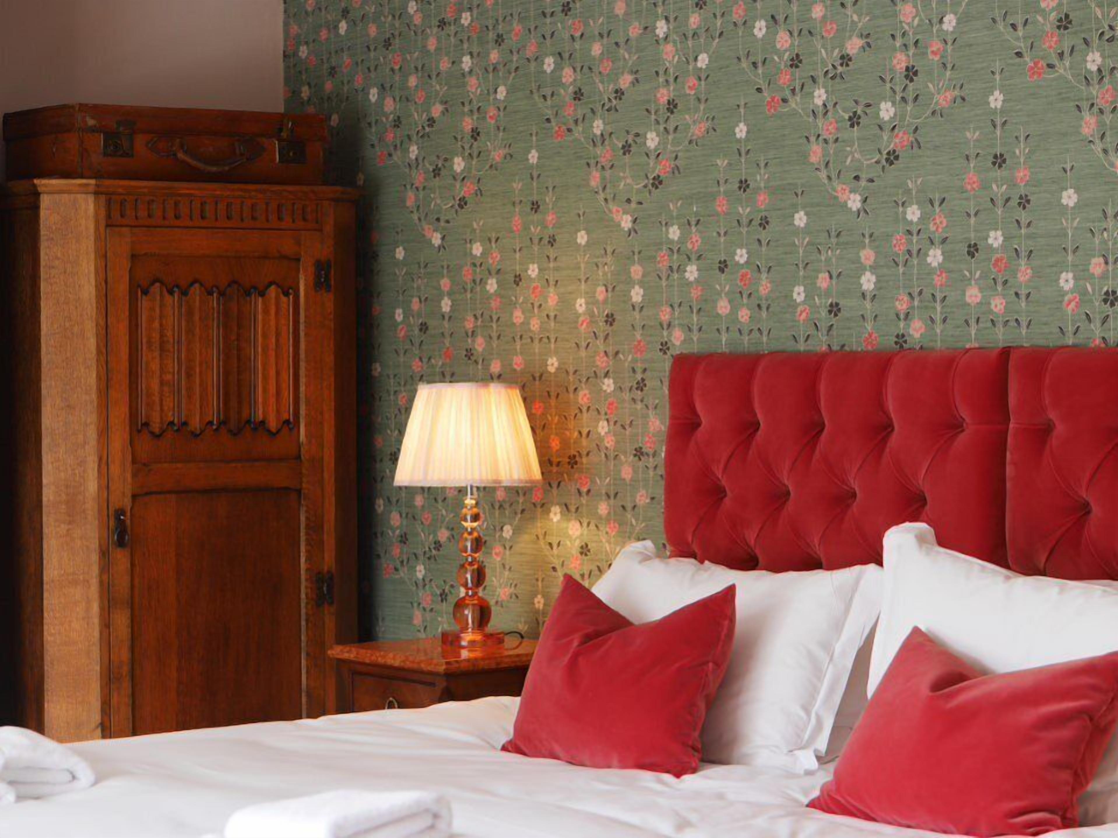 Cama bem arrumada com almofadas vermelhas e brancas, um guarda-roupa de madeira e um papel de parede floral.