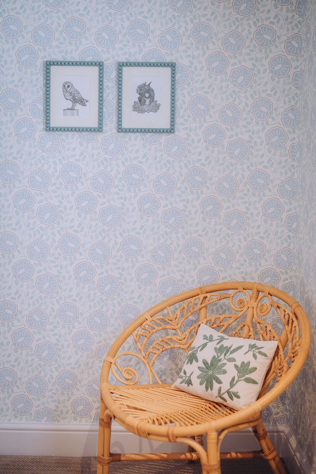 A wicker chair with a floral pillow against a wall with floral wallpaper and two framed pictures.