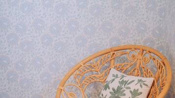 A wicker chair with a floral pillow against a wall with floral wallpaper and two framed pictures.