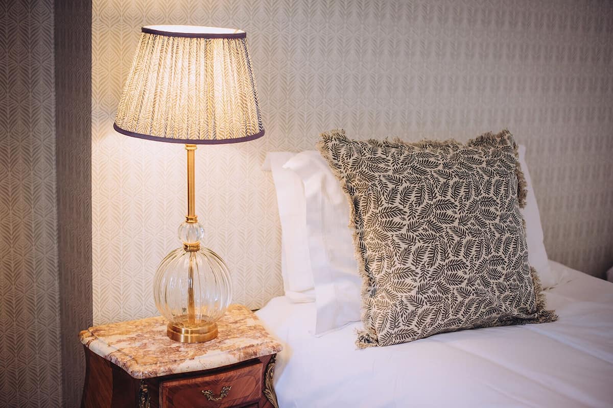 A neatly made bed with a patterned pillow, a lamp on a marble nightstand, and a patterned wallpaper.