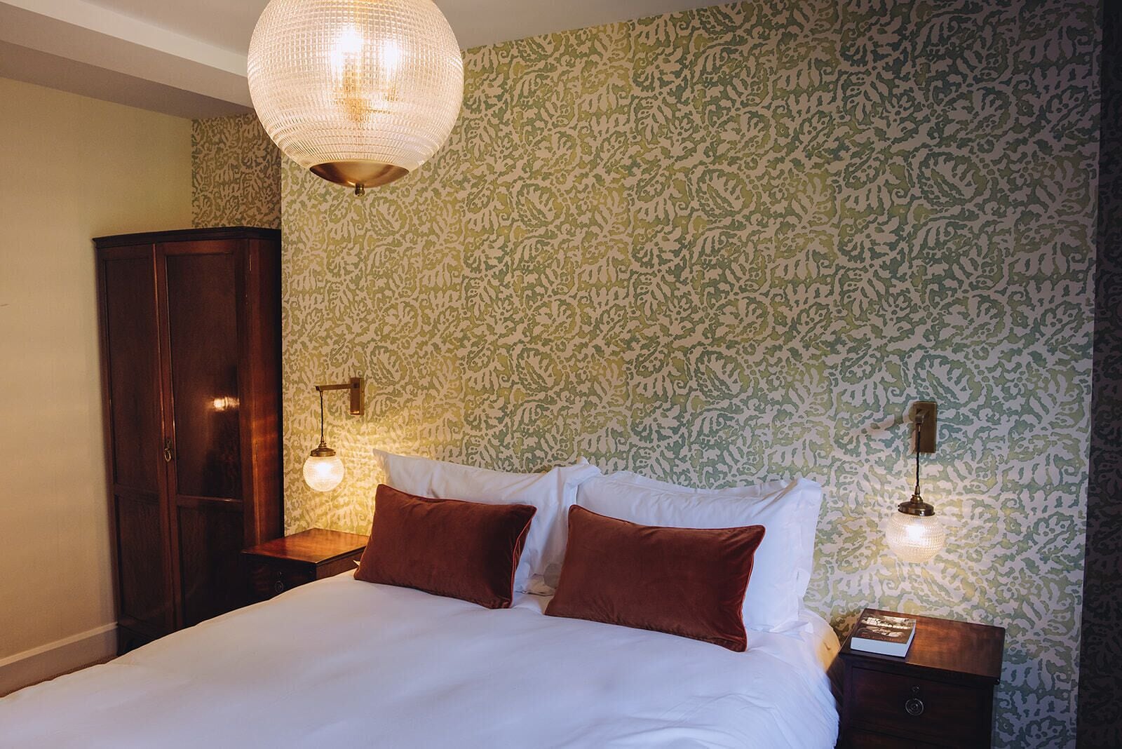 A bedroom with a large bed, two bedside tables, a wardrobe, and patterned wallpaper.