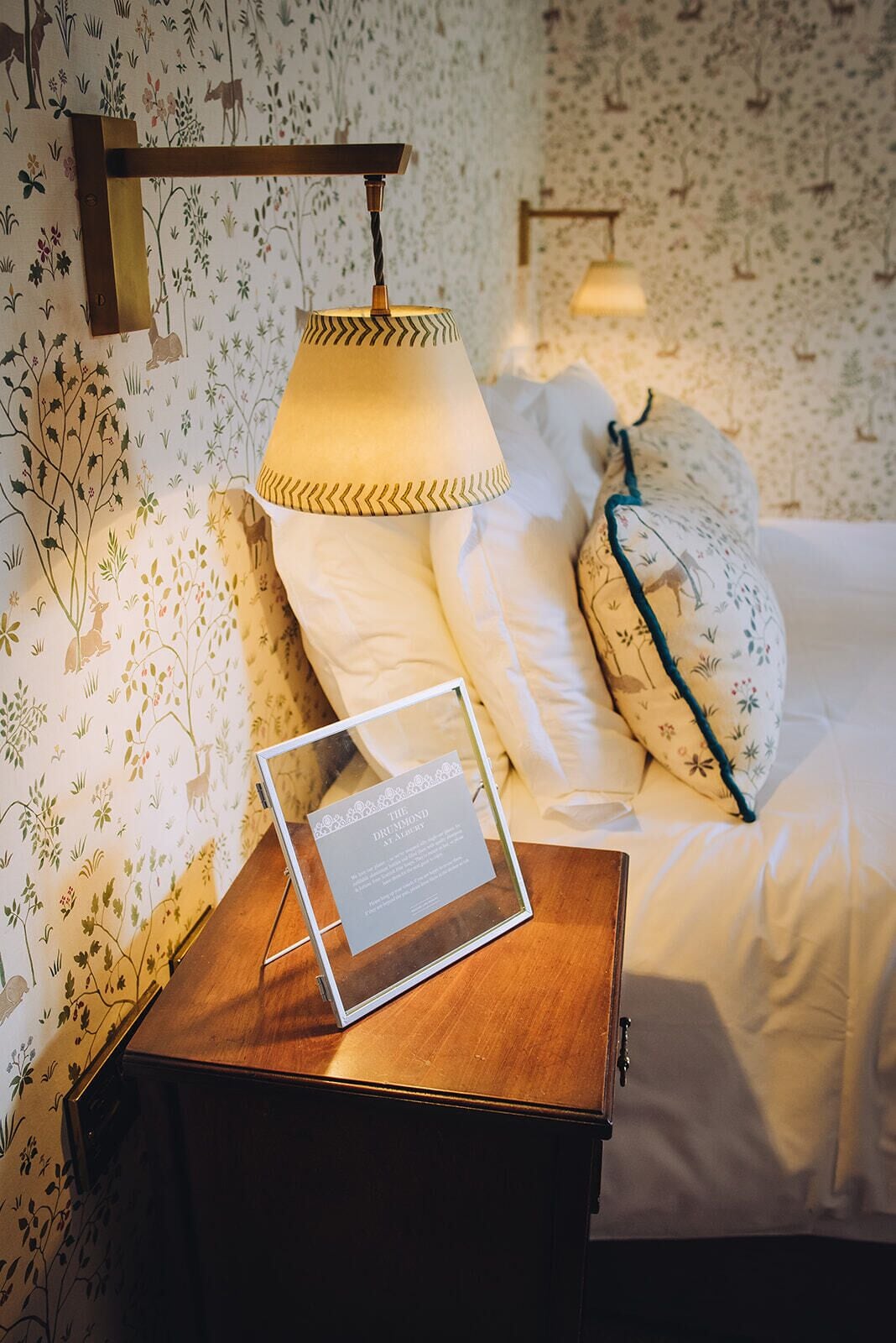 A neatly made bed with white linens and decorative pillows, a bedside table with a framed certificate, and a wall-mounted lamp.