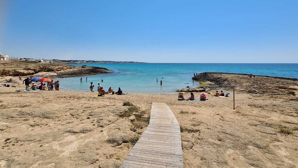 A beach with a wooden walkway leading to the water, people sunbathing and swimming, and rocky terrain. - Villetta Lorena 100mt from the sea, Salento  (Marina di Mancaversa)