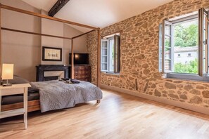 Un dormitorio con pared de piedra, una cama con cubrecama gris, una mesita de noche de madera, un televisor sobre un soporte de madera y una ventana con un persiano abierto.