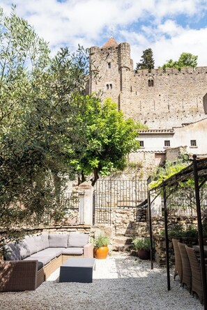 Une cour avec un mur en pierre, un grand arbre, des sièges extérieurs et, au loin, un château.