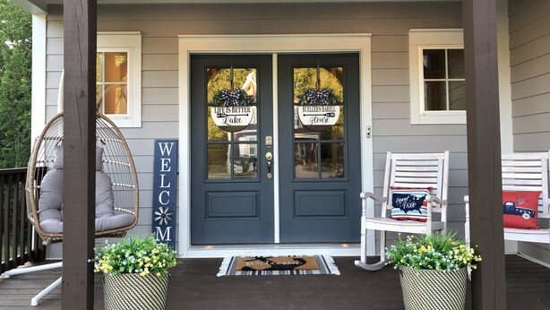 A front porch with a dark blue door, a 'WELCOME' sign, two wicker chairs, and potted plants.