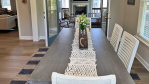 A dining area with a long table, white chairs, a chandelier, and a fireplace in the background.