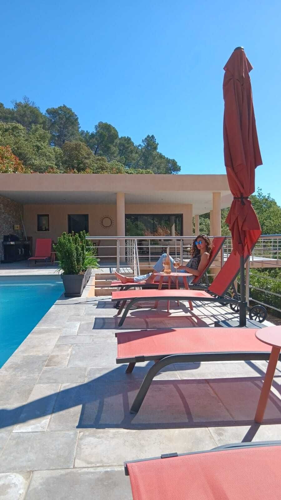 A poolside area with lounge chairs and umbrellas, a clear blue sky, and a building with a covered patio in the background.