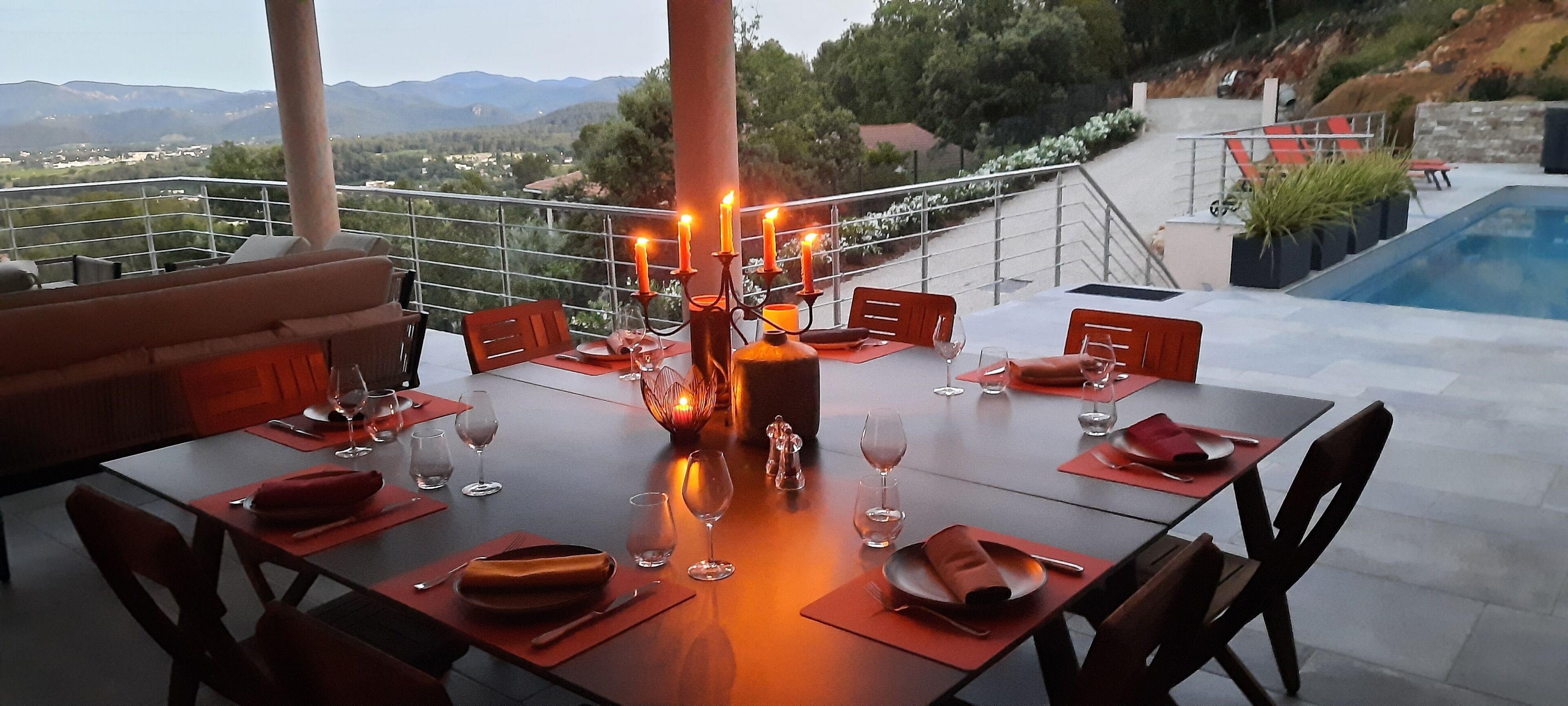 Mesa de comedor preparada para seis personas, con velas, copas de vino y platos, con vistas a una piscina y montañas.