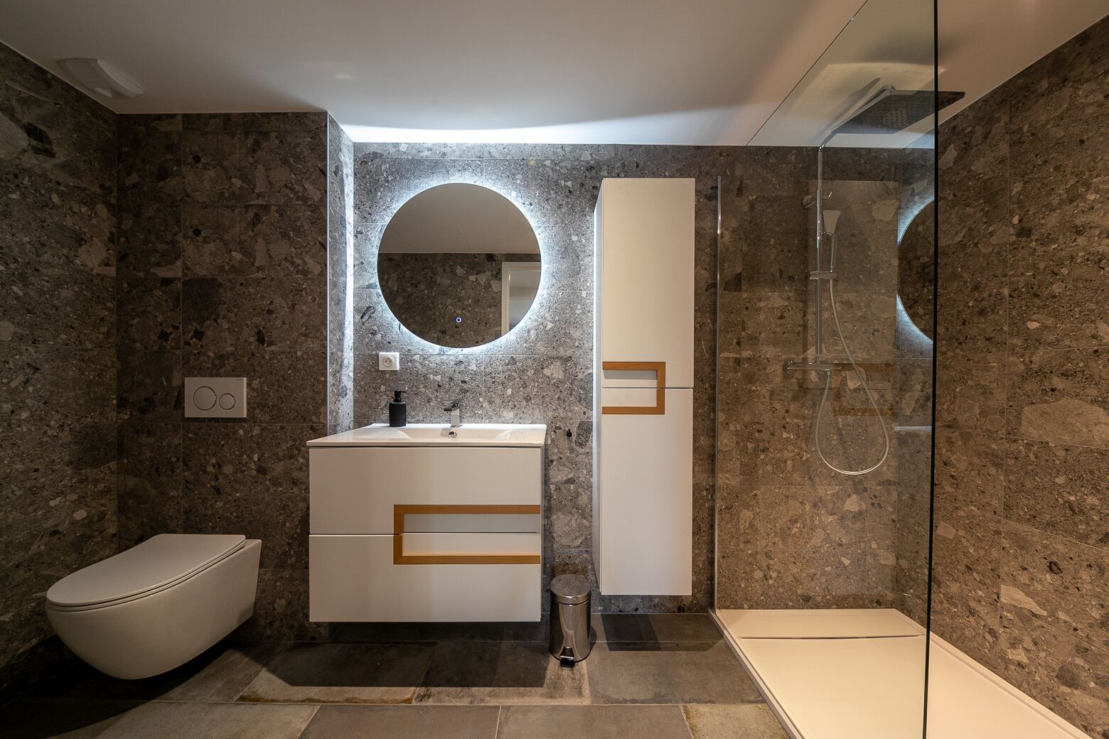 A modern bathroom with a marble wall, a round mirror, a toilet, a sink with a soap dispenser, and a glass-enclosed shower.
