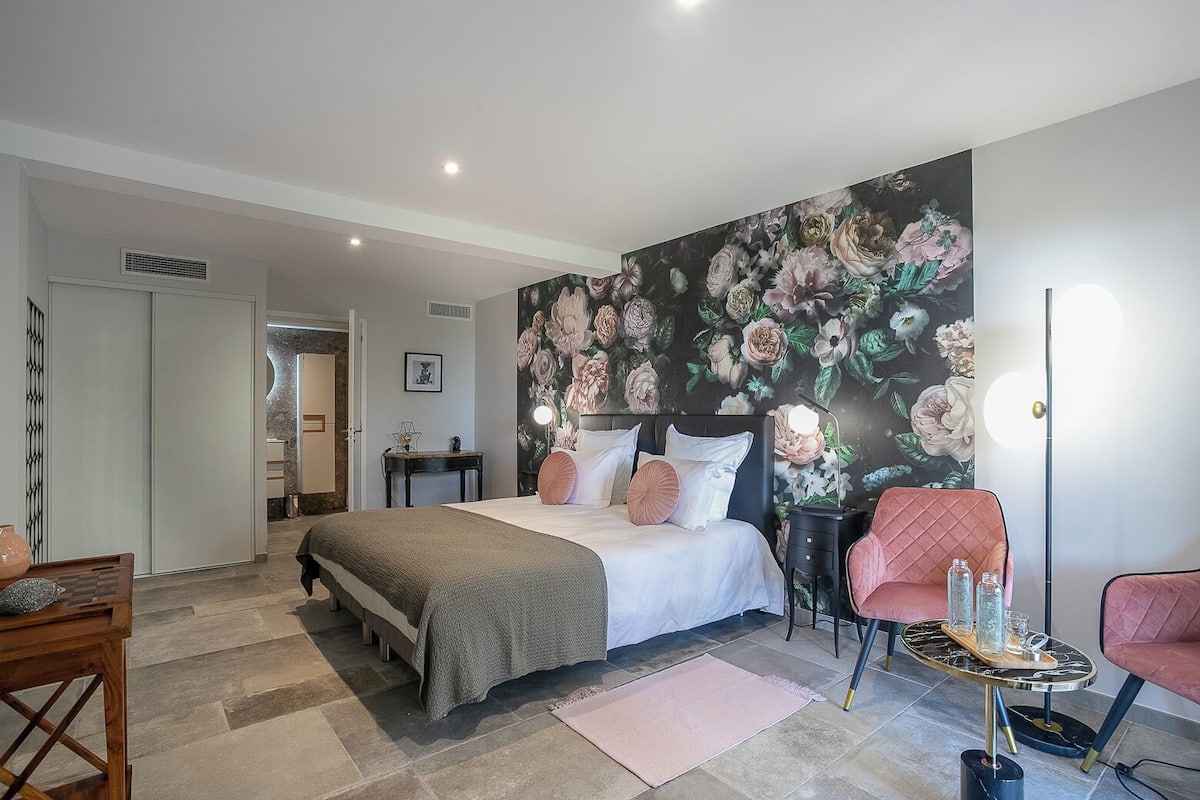 Ein Schlafzimmer mit einem Bett, einem Nachttisch, einem Sessel und einer Wand mit Blumenmuster-Tapete.