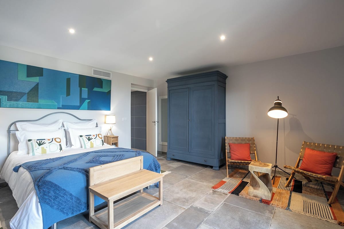 Ein Schlafzimmer mit einem Bett, einer hölzernen Bank, einem blauen Kleiderschrank, einem Sessel und einer Stehlampe.