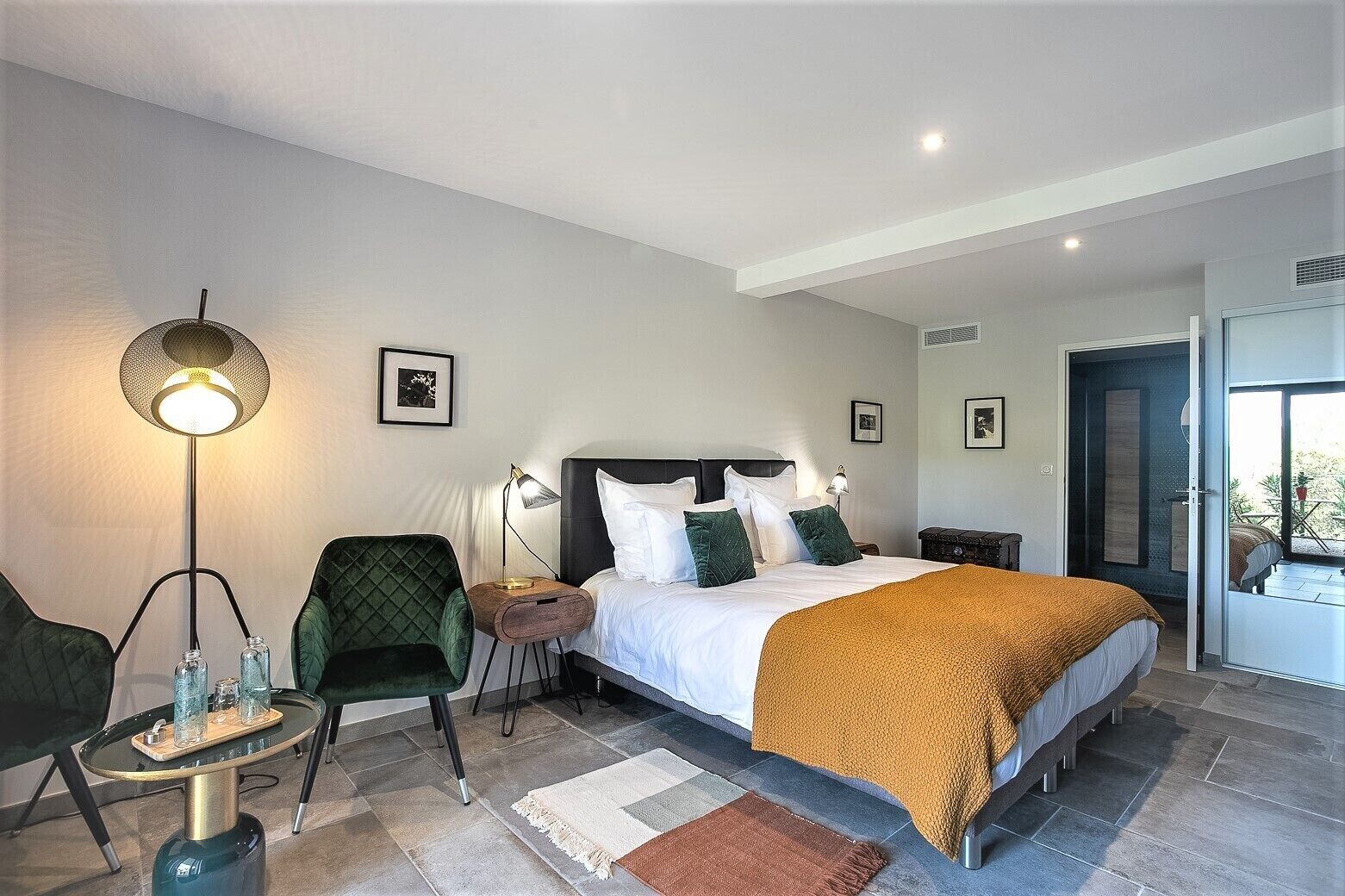 Ett modernt hotellrum med en stor säng, en grön fåtölj, ett sidobord med en lampa och utsikt över omgivningarna genom en öppen dörr.