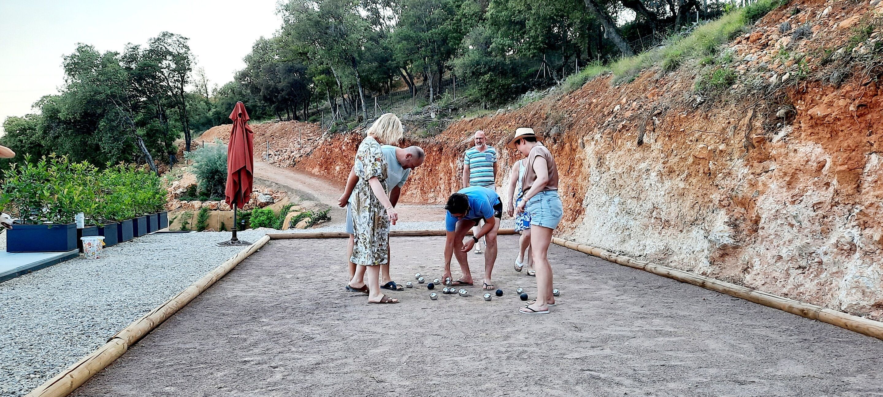Eine Gruppe von Menschen spielt ein Ballspiel auf einem Kiesplatz mit einer Erdwallung und Bäumen im Hintergrund.