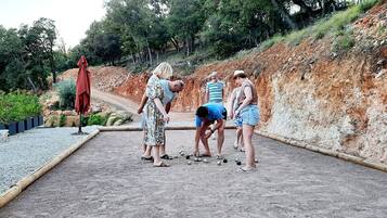 Eine Gruppe von Menschen spielt ein Ballspiel auf einem Kiesplatz mit einer Erdwallung und Bäumen im Hintergrund.