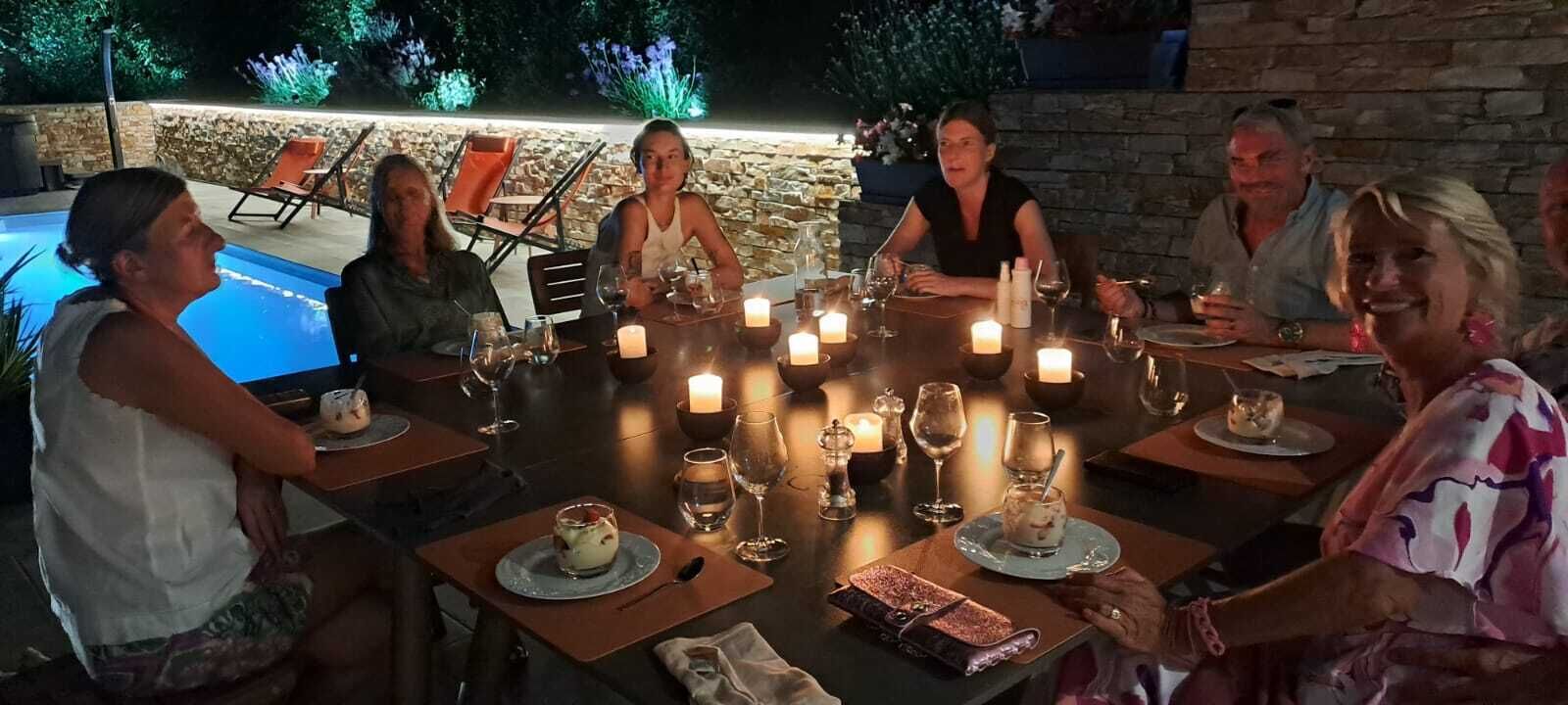A group of people dining at an outdoor table with candles, surrounded by a pool and stone walls.