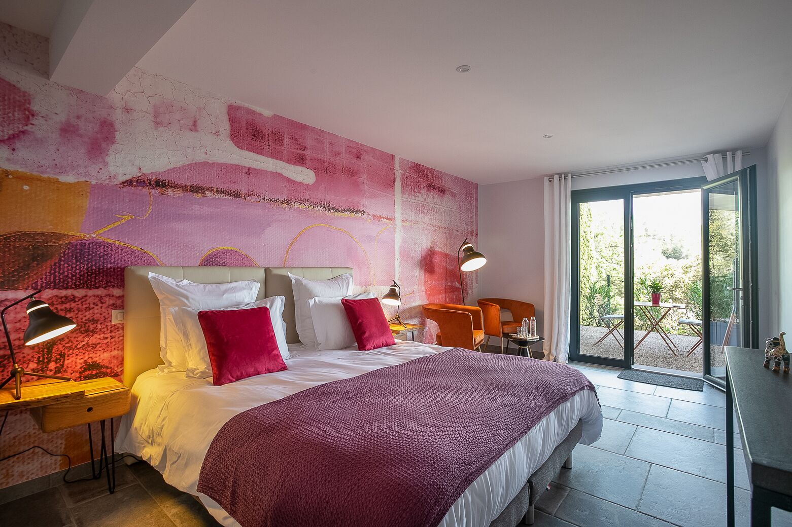 A modern bedroom with a large bed, two bedside tables with lamps, a view of the outdoors through a sliding glass door, and a vibrant abstract wall mural.