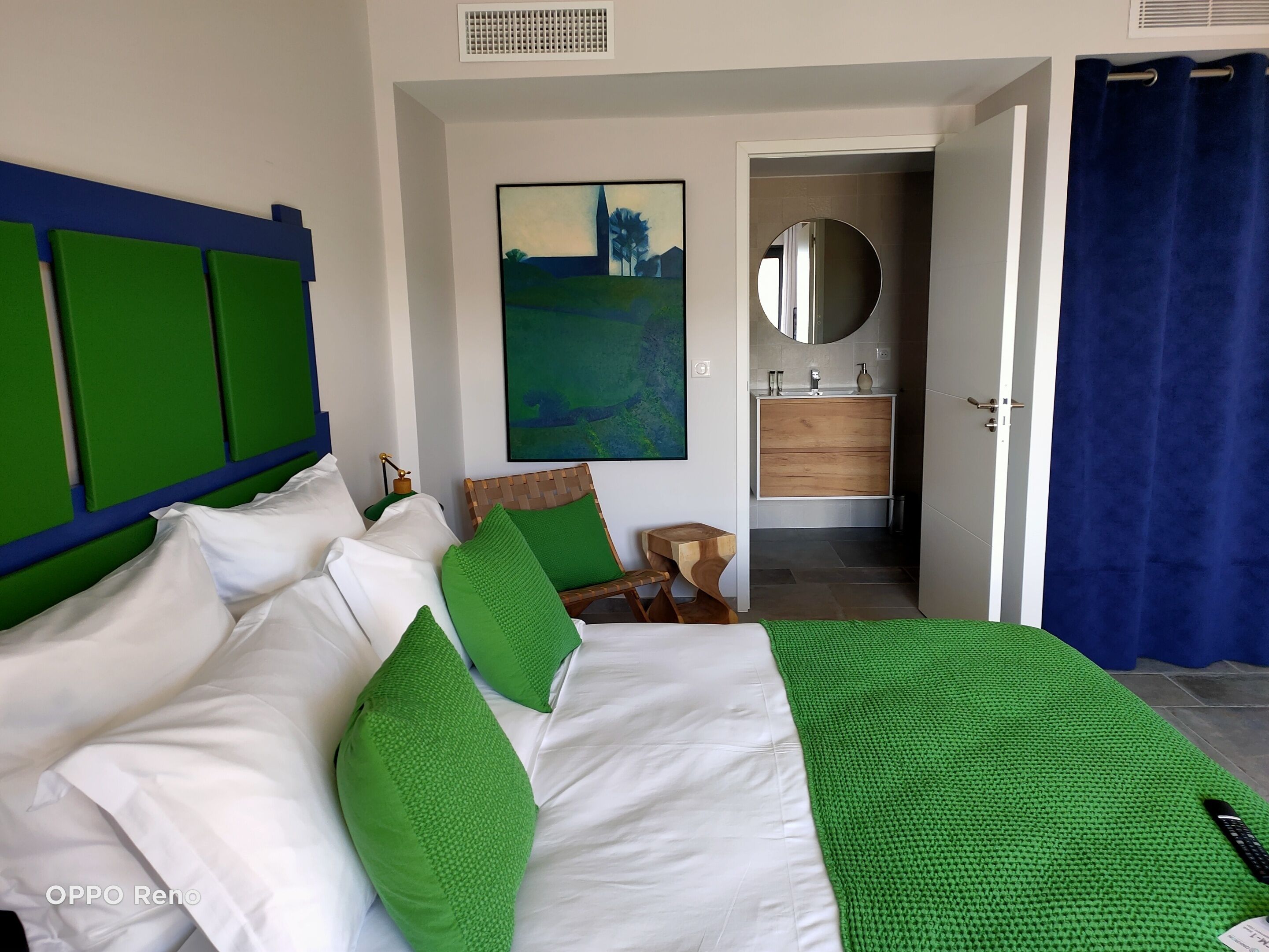 Une chambre à coucher avec un lit, des oreillers verts, une tête de lit en bois, un tableau, un miroir, une armoire en bois et un rideau bleu.