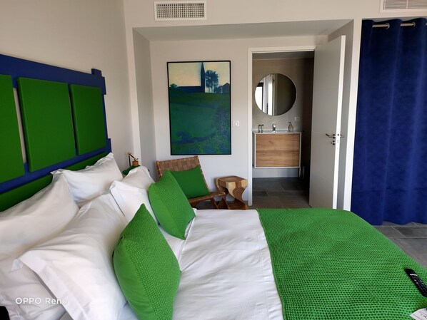 Un dormitorio con cama, almohadas verdes, cabecera de madera, un cuadro, un espejo, un armario de madera y una cortina azul.