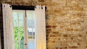 Une chambre avec un mur en briques, un plancher en bois, un lit avec une literie à motifs, une table de chevet avec une lampe et une fenêtre avec des rideaux.