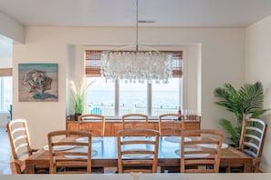 A dining area with a wooden table and chairs, a chandelier, and a view of the sea through a window with blinds.