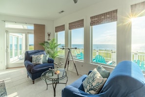 A spacious living room with a large window overlooking the sea, furnished with a blue sofa, a glass coffee table, and a tripod.