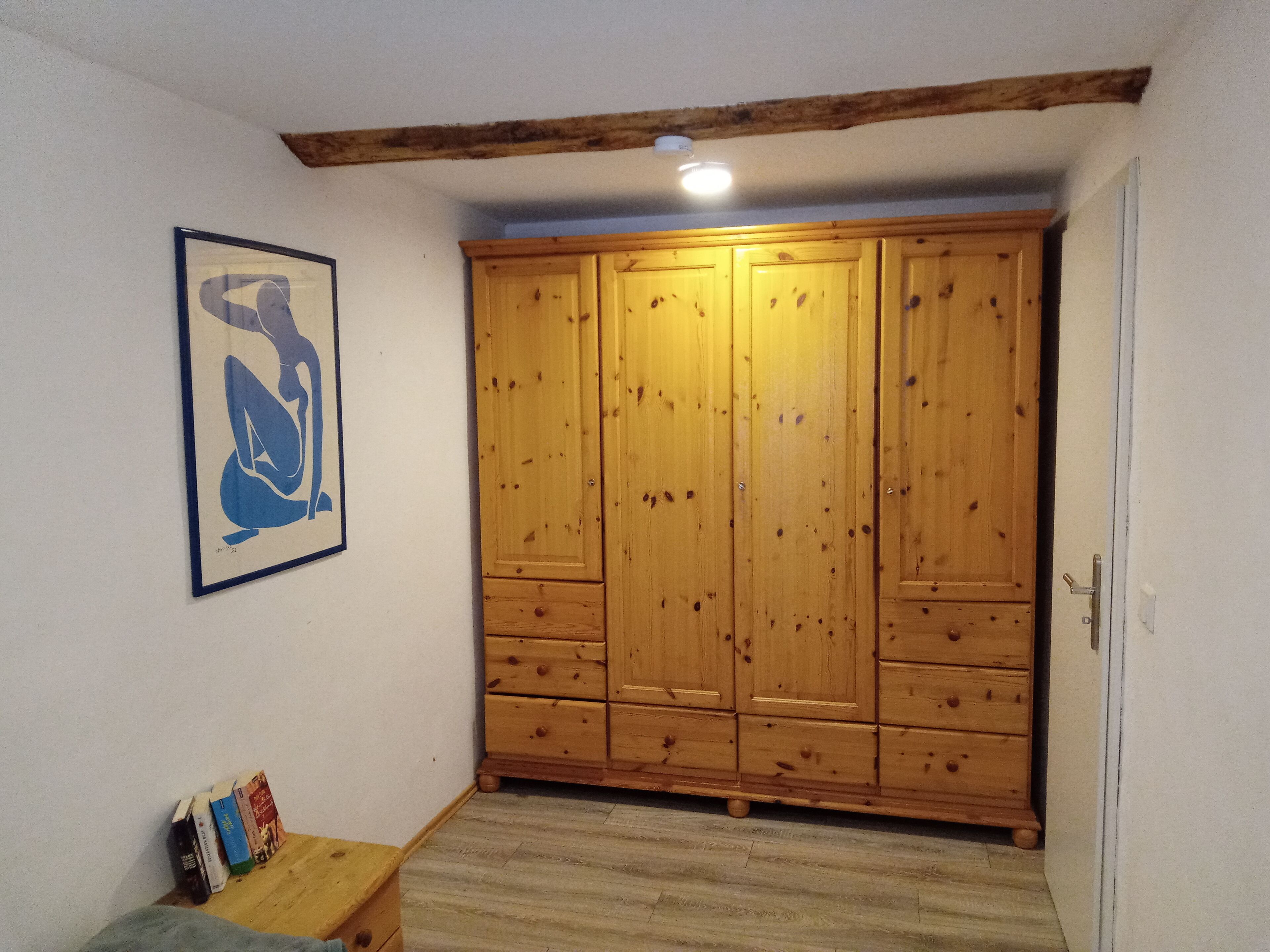 Ein Zimmer mit einem Holzschrank, einem gerahmten Kunstwerk an der Wand und einem kleinen Holztisch mit Büchern.