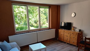 A room with a bed, a small white table, a TV on a wooden dresser, and a window with a view of greenery.