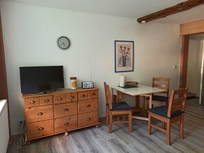 Ein kleiner, gemütlicher Raum mit einem Holzkonsolenmöbel, einem Fernseher, einer Uhr, einem gerahmten Bild und einem Essbereich mit Stühlen und einem Tisch.