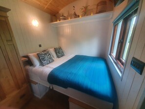 A compact bedroom with a single bed, a nightstand, a shelf with decorative items, and a window with blinds.