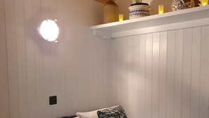A bedroom with a wooden ceiling, white walls, a bed with white and blue bedding, and a shelf with decorative items and candles. - Luxury romantic shepherd's hut - hot tub and Aga  (Camelford)