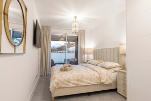 A bedroom with a bed, a bedside table, a lamp, a TV, a mirror, and a view of the outdoors. - Cala Serena, Elegant 2 bedroom apartment only 850 m from the beach in La Cala de Mijas (La Cala de Mijas)