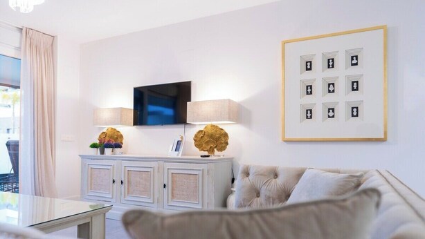 Un saló modern amb un televisor de pantalla plana penjat a la paret, un moble blanc amb objectes decoratius, un sofà amb quilles i una imatge emmarcada de cartes de joc.