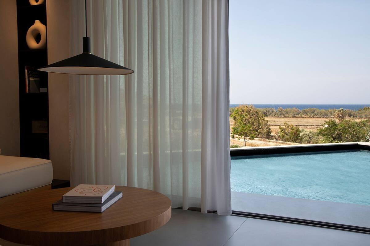 A modern room with a view of a swimming pool, a round wooden table with a book, and a black pendant light.