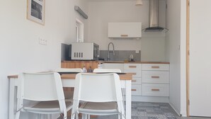 Cocina compacta con armarios blancos, un microondas y un lavamanos. Hay dos sillas blancas y una pequeña mesa. Se ve una puerta a la derecha.