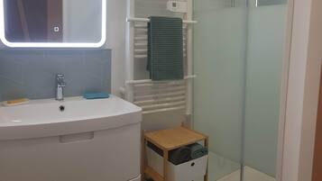 Un baño con lavamanos blanco, espejo, toallero, ducha y un pequeño mueble de almacenamiento de madera.