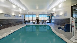 Piscina interior con señalización clara, una silla de socorrista y una zona junto a la piscina con sillas y una mesa.