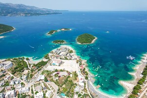 Vista aérea de una zona costera con varias islas, aguas azul turquesa cristalinas y una playa urbana con edificios y automóviles estacionados.