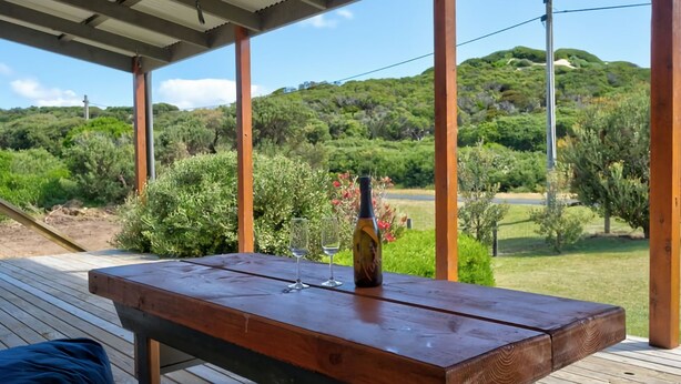 A wooden table with a bottle and glasses on a deck with a view of a hilly landscape.