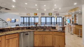 A modern kitchen with a curved island, stainless steel appliances, wooden cabinets, and large windows offering ocean views. - Penthouse on the Beach! Beautiful Sunset Views! (Fort Myers)